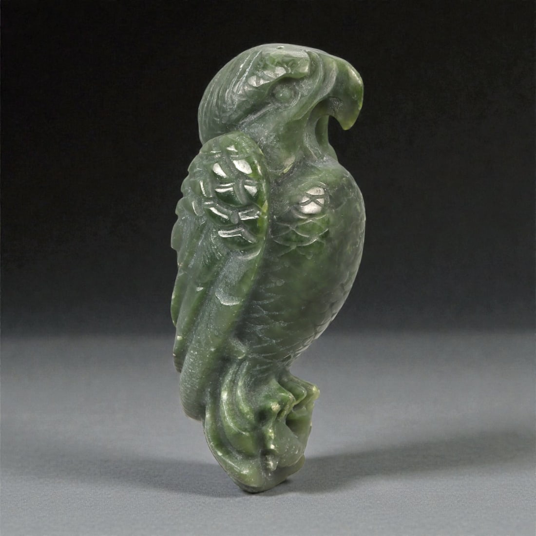 A Chinese Jade Parrot Figure, 20th Century, 23YA: Carved from dark green jade with lighter translucent areas and natural tonal variation, the figure is worked fully in the round as a standing parrot shown perched upright. The head is turned slightly