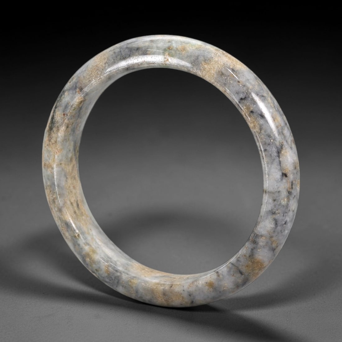 A Chinese Jadeite Jade Bangle Bracelet, 20th Century, 93ZK: Carved from jadeite jade with a pale greyish-white base and irregular tan and dark grey mottling, the bangle is formed as a continuous circular band with a rounded cross-section. The surface is smooth