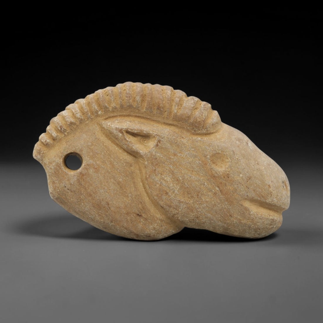 Ancient Stone Ibex Amulet - Near Eastern Carved Figurine, 10ZX: Carved from light-colored hardstone, the amulet takes the form of a stylized ibex head, rendered in three dimensions with detailed relief carving. A suspension hole is drilled through the rear of the