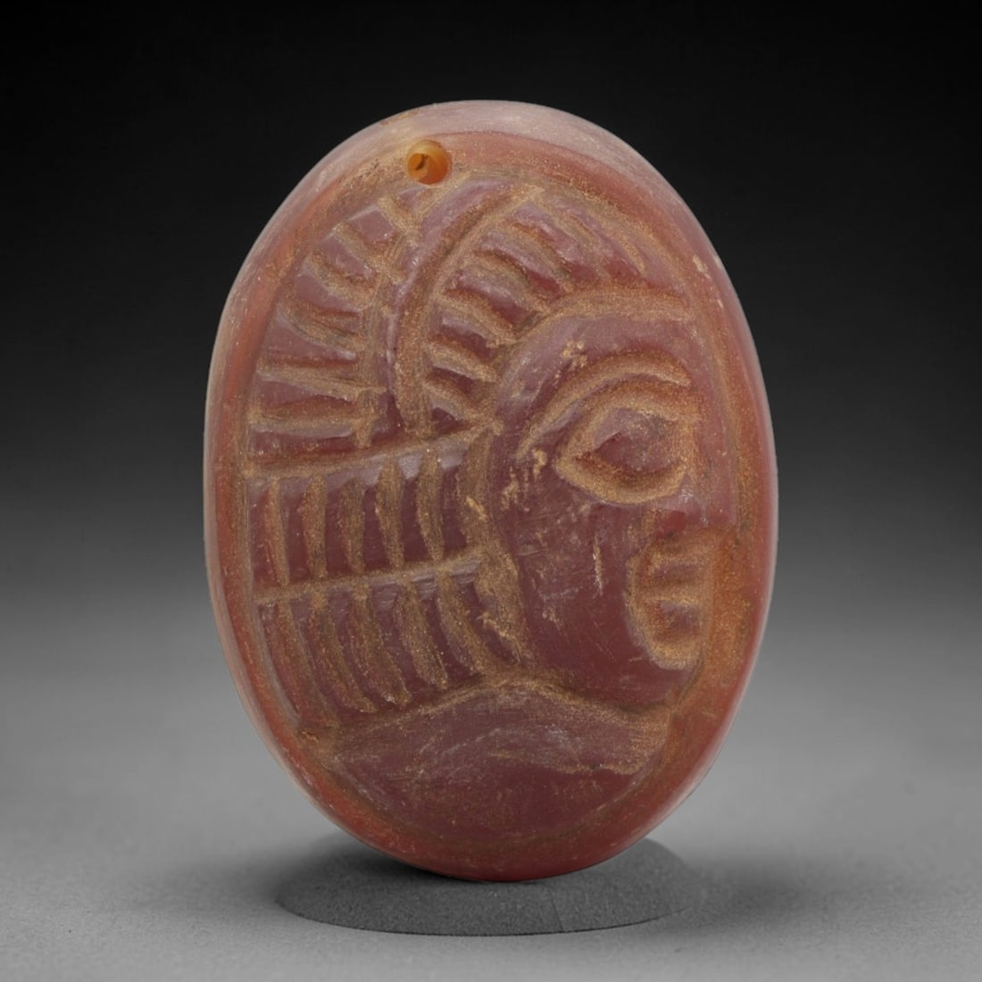 Ancient Carnelian Amulet - Near Eastern Bust Pendant, 50ZX: Carved from carnelian, the amulet is an oval disc with a drilled suspension hole at the top, crafted in the ancient Near Eastern style. The front surface bears a raised relief portrait of a human bust