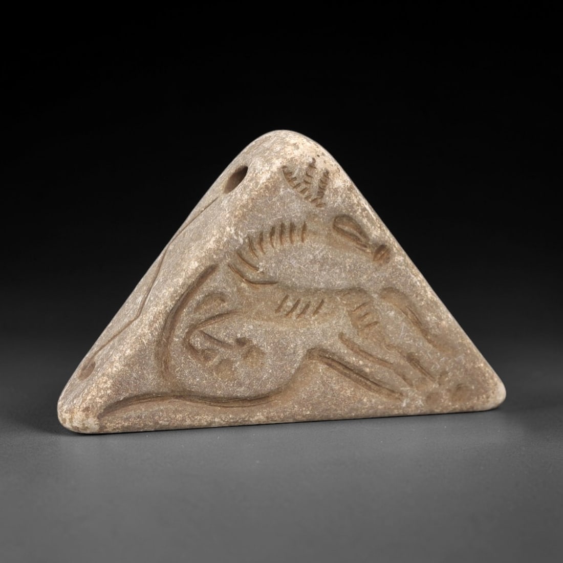 Ancient Stone Double Convex Amulet - Near Eastern Ibex Figurine, 14FX: Carved from smooth, pale stone, this double convex triangular amulet features a low-relief depiction of a rearing ibex, with incised linear details defining the horns, body, and limbs, alongside a sma