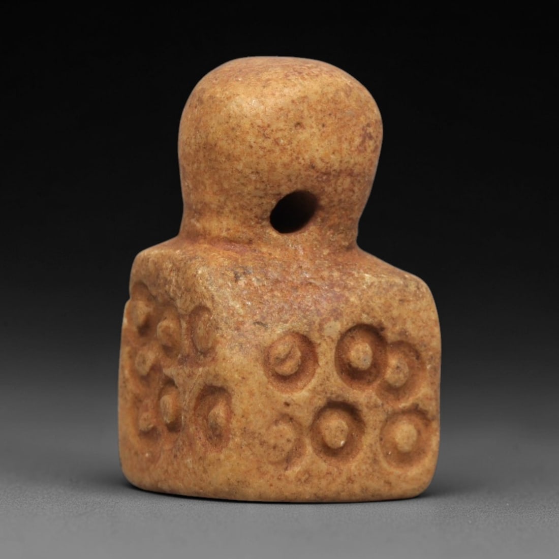 Ancient Stone Amulet - Near Eastern Concentric Circles, 55ZP: This amulet is carved from granular stone, featuring a rounded upper section with a suspension hole and a square lower body decorated with incised concentric circle motifs arranged in clusters. Stylis