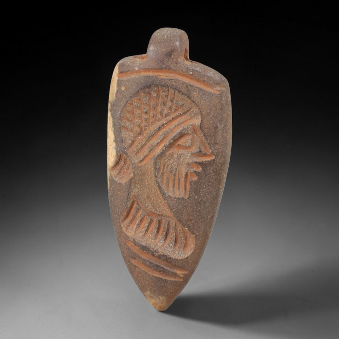 Ancient Stone Head Amulet - Near Eastern Plaque, 30FY: Carved from a dense, dark brown stone with warm mineral inclusions, this amulet takes the form of an elongated, leaf-shaped plaque with a small suspension lug at the top, characteristic of ancient Nea