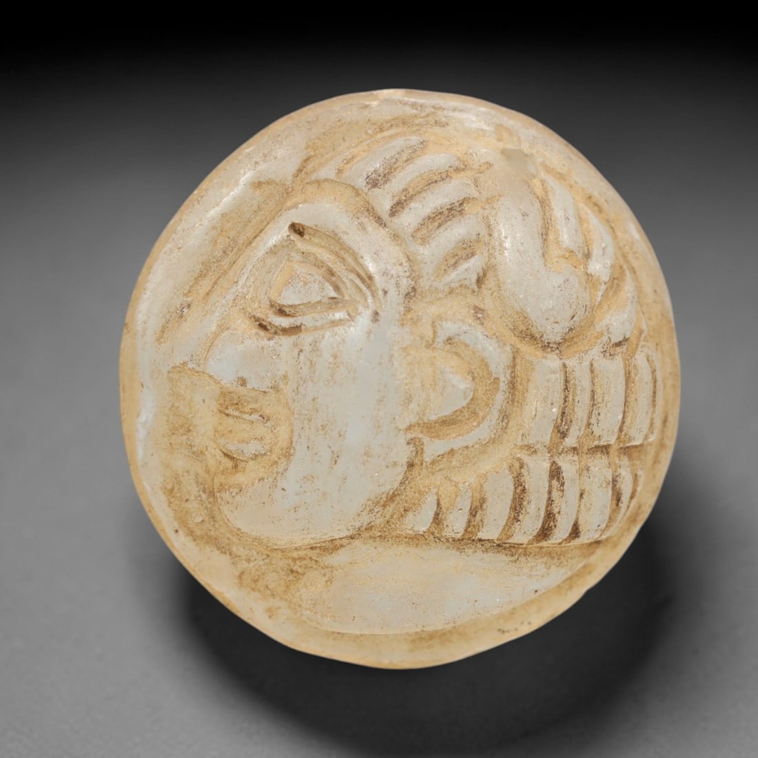 Ancient Glass Medieval Amulet - Near Eastern Bust Figure, 73ZR: Carved from translucent pale glass with earthy mineral encrustations, this spherical amulet showcases a raised relief profile bust of a human figure, characterized by defined facial features, a styliz