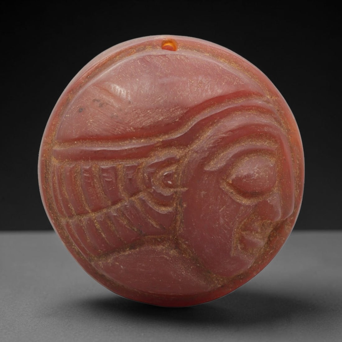 Ancient Carnelian Amulet - Near Eastern Bust Pendant, 42ZX: Carved from carnelian, the amulet is a circular disc with a drilled suspension hole at the top edge, crafted in the ancient Near Eastern style. The front surface bears a raised relief portrait of a hu