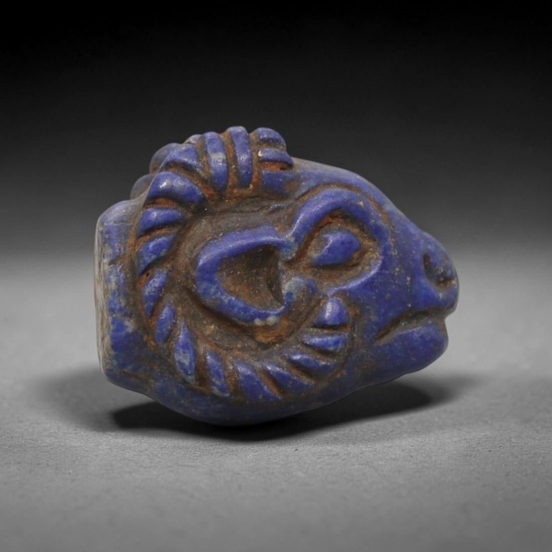 Ancient Lapis Lazuli Ram Head - Near Eastern Amulet Figurine, 99XV: Carved from lapis lazuli, this amulet depicts a stylized ram head with prominent, spiraled horns and detailed facial features. The eyes are deeply incised, the snout is defined, and the horns are rend