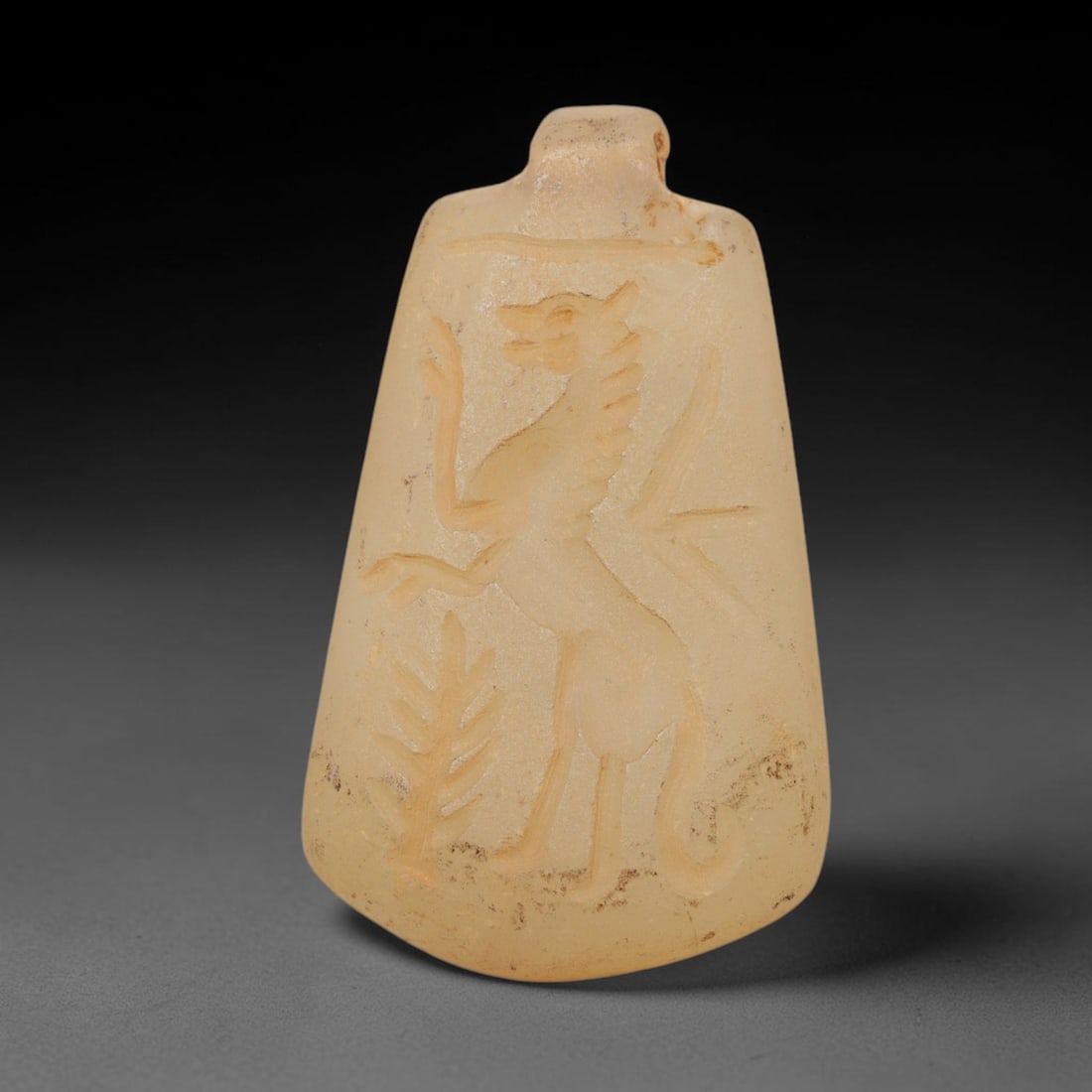 Ancient Chalcedony Amulet - Near Eastern Griffin Figure, 48ZX: Carved from translucent chalcedony, this amulet features a relief depiction of a standing griffin, a mythical hybrid creature, rendered in a stylized Near Eastern glyptic tradition. The figure is show