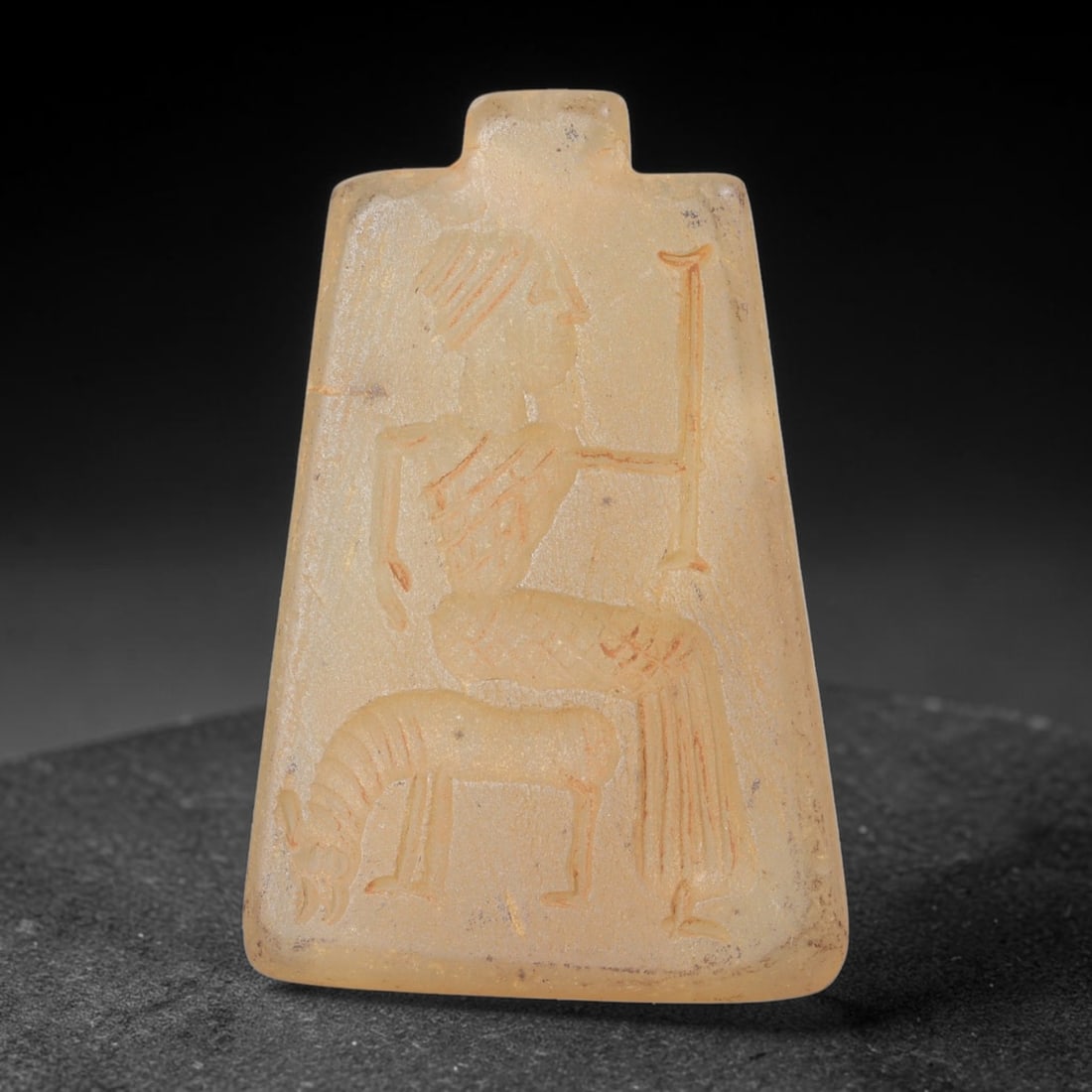 Ancient Chalcedony Figure Amulet - Near Eastern Plaque, 26FY: Carved from a pale, translucent chalcedony, this amulet takes the form of a trapezoidal plaque with a small suspension lug at the top, characteristic of ancient Near Eastern amuletic and glyptic tradi