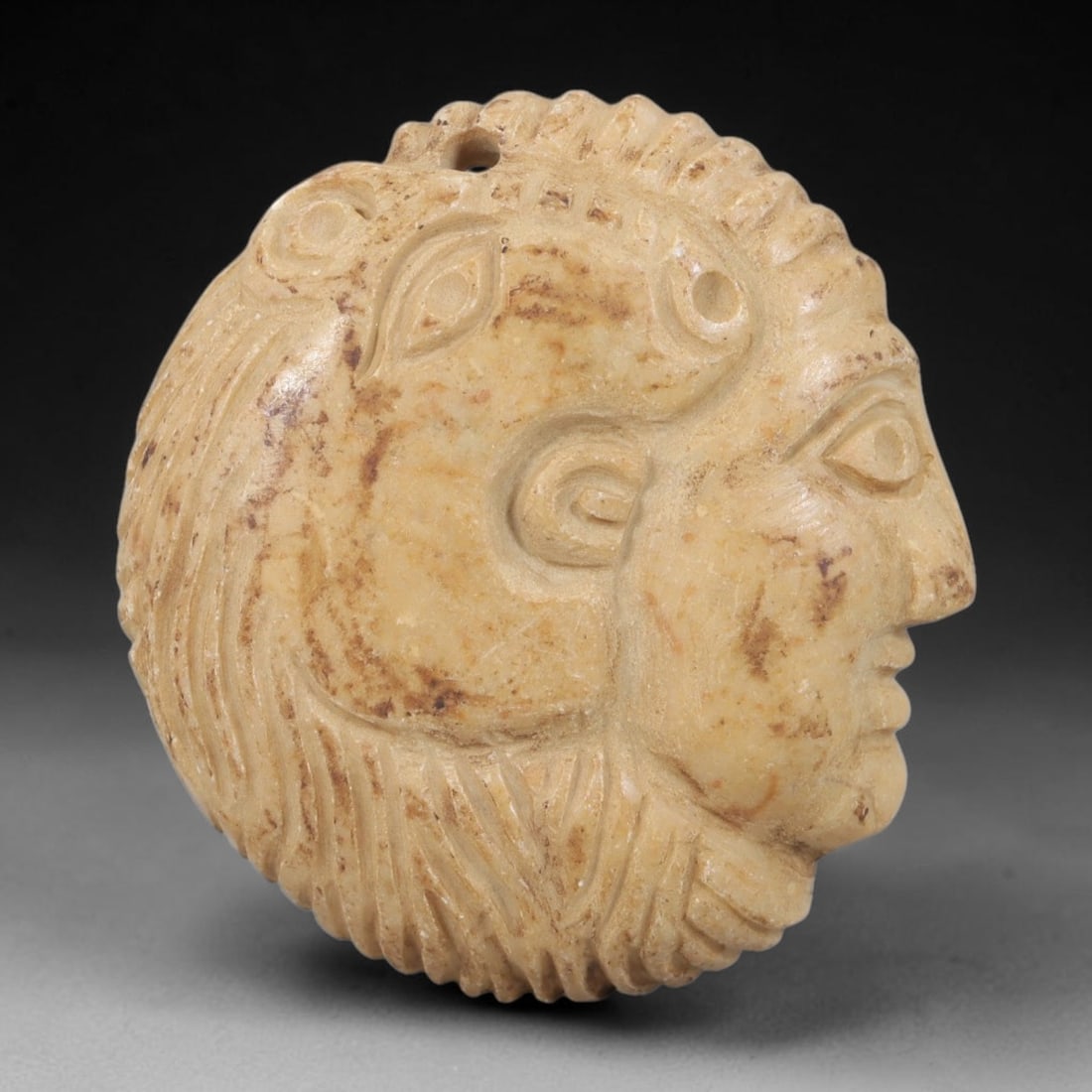 Ancient Stone Amulet - Near Eastern Ram-Headed Deity Pendant, 34FY: Carved from light-colored stone, the amulet is a circular pendant crafted in the ancient Near Eastern style, featuring a raised relief portrait of a ram-headed deity in profile. The surface bears inci