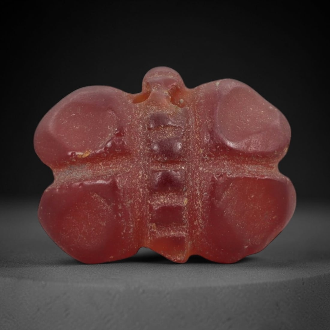 Ancient Carnelian Butterfly Amulet - Egyptian Figurine, 52AJ: Carved from deep red carnelian, this amulet is formed as a stylized butterfly with four rounded wing lobes and a segmented central body, featuring incised detailing along the torso to denote anatomica