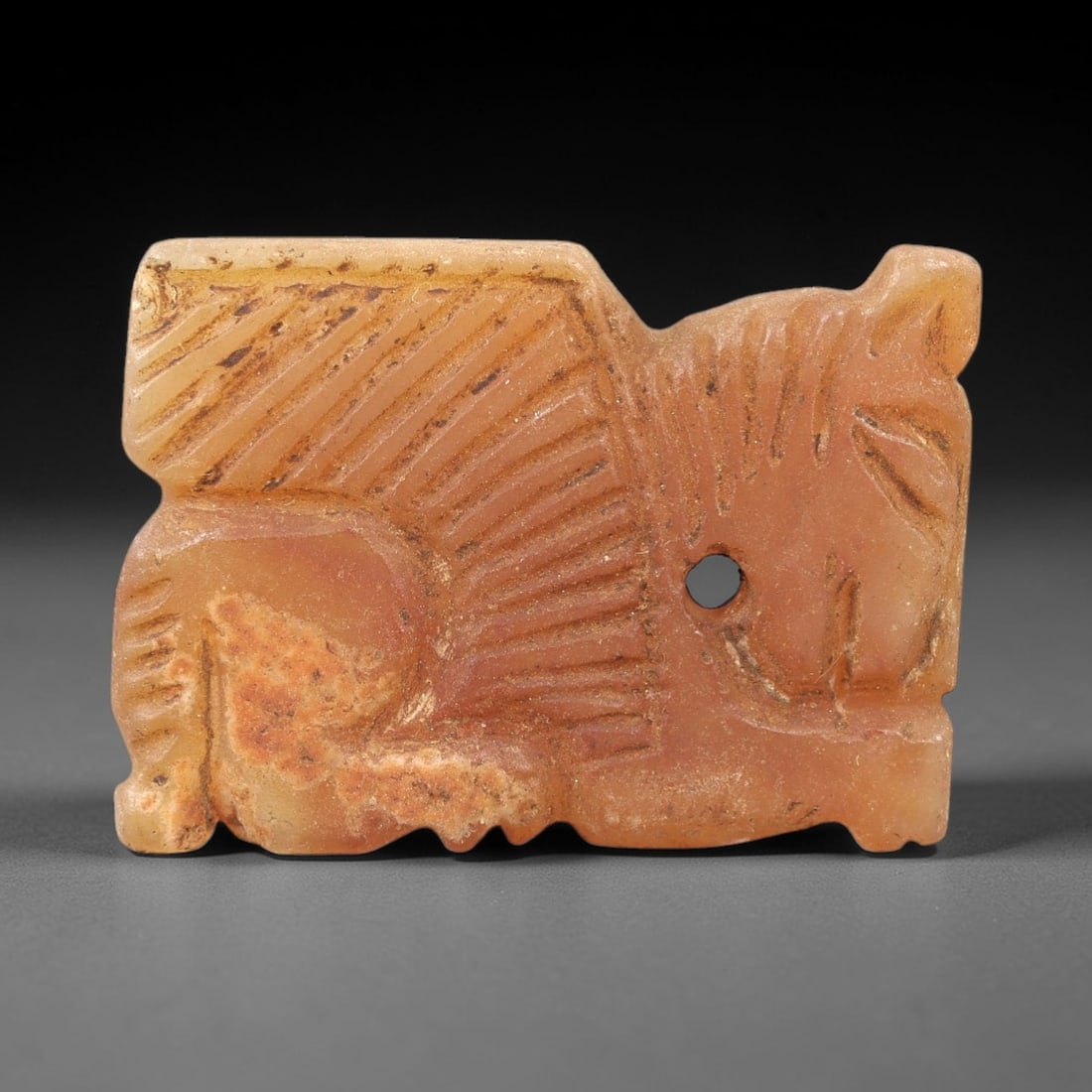 Ancient Chalcedony Lamassu - Near Eastern Amulet Pendant, 26AK: Carved from warm orange chalcedony, this miniature amulet depicts a stylized lamassu (winged guardian beast) in incised relief, with linear patterns defining its feathered wings, striped mane, and cro