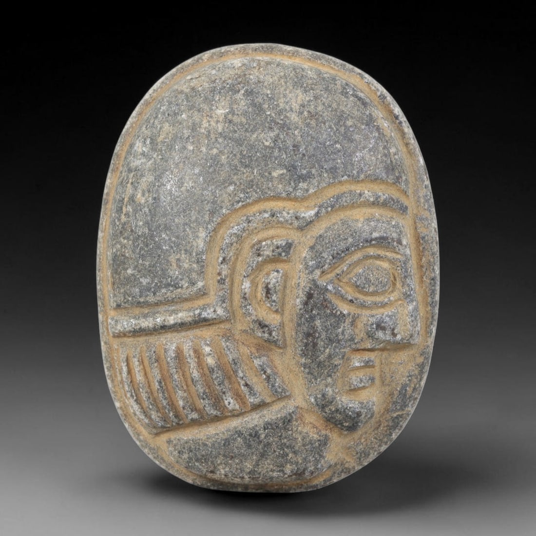 Ancient Stone Head Plaque - Near Eastern Amulet, 35FY: Carved from a dense, dark grey stone with a weathered surface, this amulet takes the form of a stylized human head in high relief, set within an oval plaque, characteristic of ancient Near Eastern vot