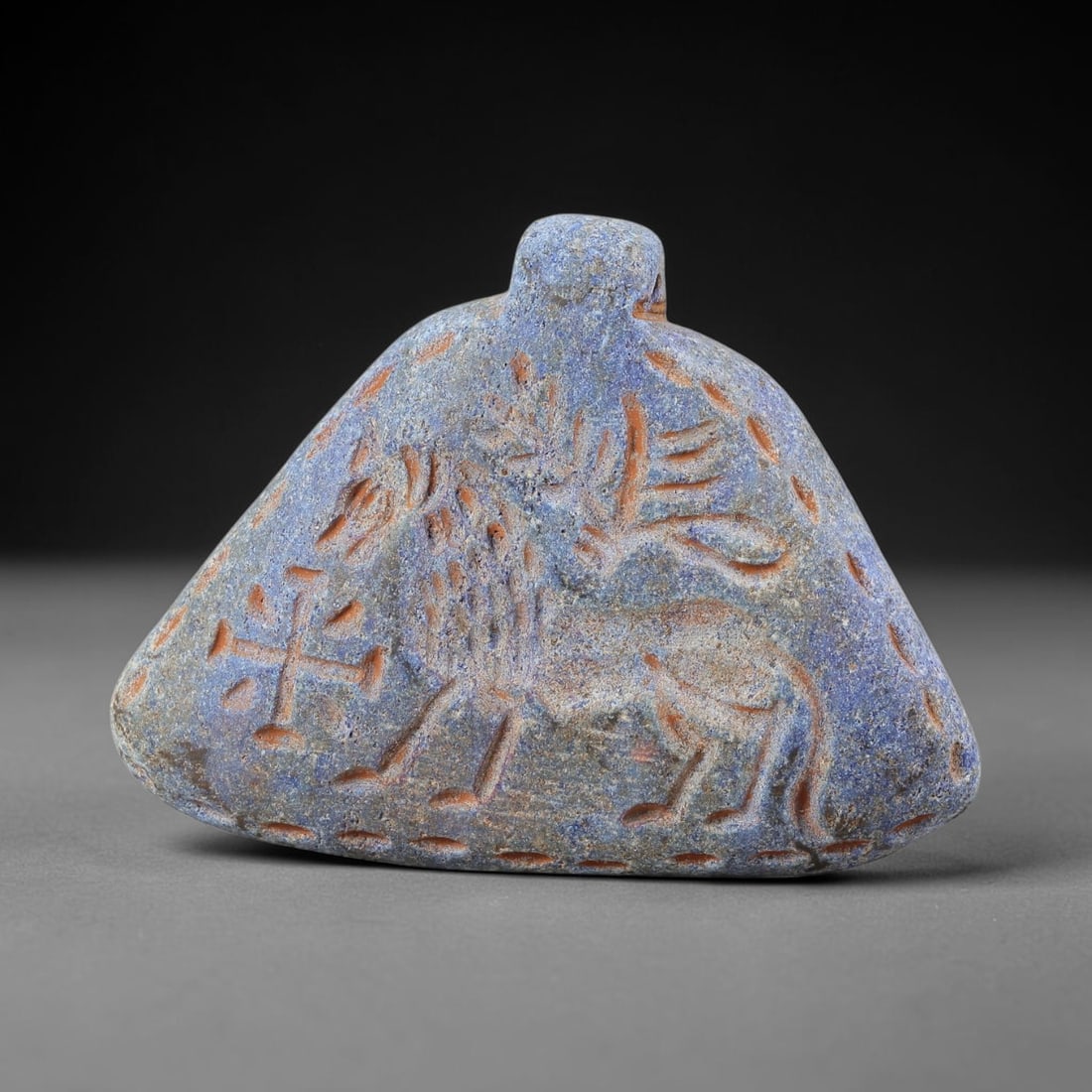 Ancient Lapis Lazuli Amulet - Near Eastern Stag Figurine, 30BA: Carved from lapis lazuli, the amulet takes a triangular, bell-shaped form with a suspension loop at the top, designed for wear as a protective talisman. The front face is incised with a stylized stag