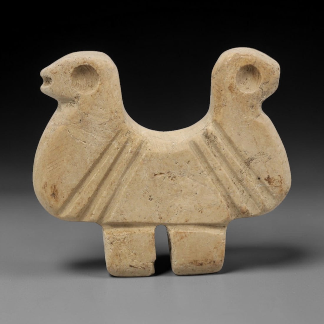 Ancient Limestone Double-Headed Animal - Near Eastern Figurine, 60ZW: This object is carved from pale beige limestone, depicting a double-headed animal with two stylized heads facing opposite directions, each with a circular perforation for suspension. The body is geome