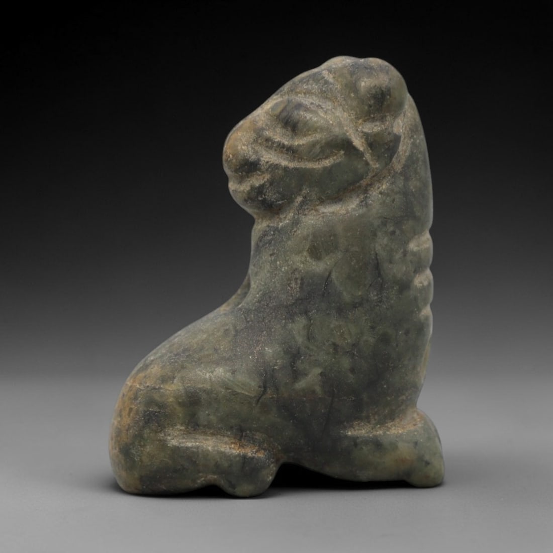Ancient Near Eastern Stone Ram Amulet - Carved Figurine, 93ZW: This amulet is carved from a mottled, greenish-grey stone, likely serpentine or steatite, with incised linear details defining its stylized recumbent ram form. The piece depicts a ram in a seated pose