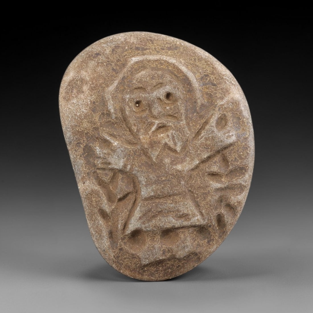 Ancient Stone Amulet - Near Eastern Christian Saint, 10FY: Carved from a naturally smoothed river stone, this amulet features a bas-relief depiction of a bearded male saint, identifiable by the incised nimbus or halo framing his head. The figure is rendered w