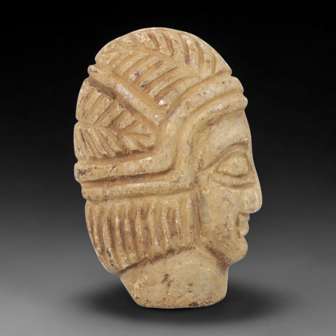 Ancient Stone Head - Near Eastern Anthropomorphic Figurine, 37FY: Carved from light-colored stone, the artifact is a three-dimensional human head in profile, crafted in the ancient Near Eastern style. The surface bears incised details rendering an elaborate, chevron