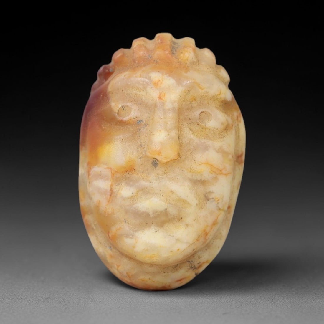 Ancient Stone Anthropomorphic Head Amulet - Near Eastern Ritual Figurine, 29AG: This amulet is carved from a single piece of mottled pale orange and cream stone, exhibiting a smooth, semi-translucent texture with subtle ochre veining and natural mineral inclusions across its surf