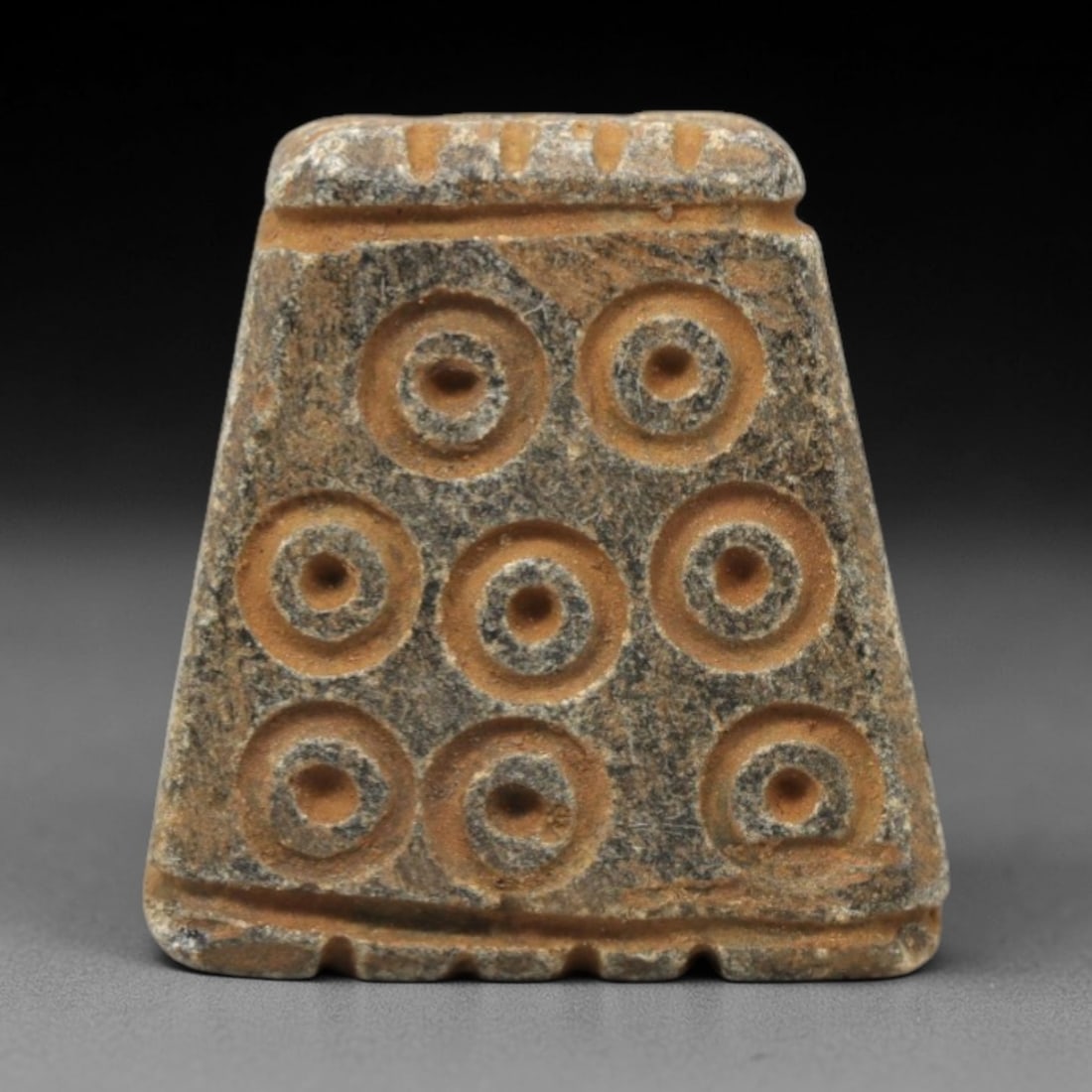 Ancient Steatite Amulet - Near Eastern Concentric Circles, 36XV: Carved from compact steatite, this ancient amulet features a trapezoidal form, adorned with a grid of eight concentric circle motifs, each with a central dot, executed with precise, incised carving ty