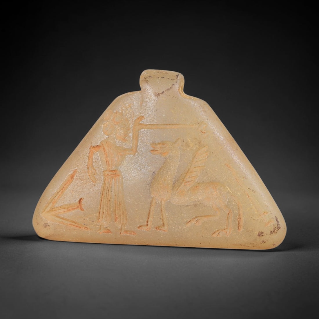 Ancient Stone Master of Animals Amulet - Near Eastern Carved Pendant, 19BA: Carved from translucent light-colored hardstone, the triangular amulet features a relief-carved figural scene, rendered with incised detail and filled with red pigment for contrast. The composition de