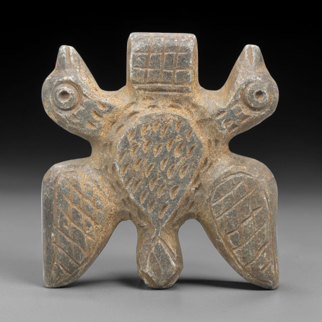Ancient Near Eastern Double Bird Amulet - Figurine, 40ZR: This artifact is a carved stone zoomorphic amulet, crafted from a dense, grey-brown stone with a textured, matte surface and natural earthy patina. The piece takes the form of a symmetrical double-bir