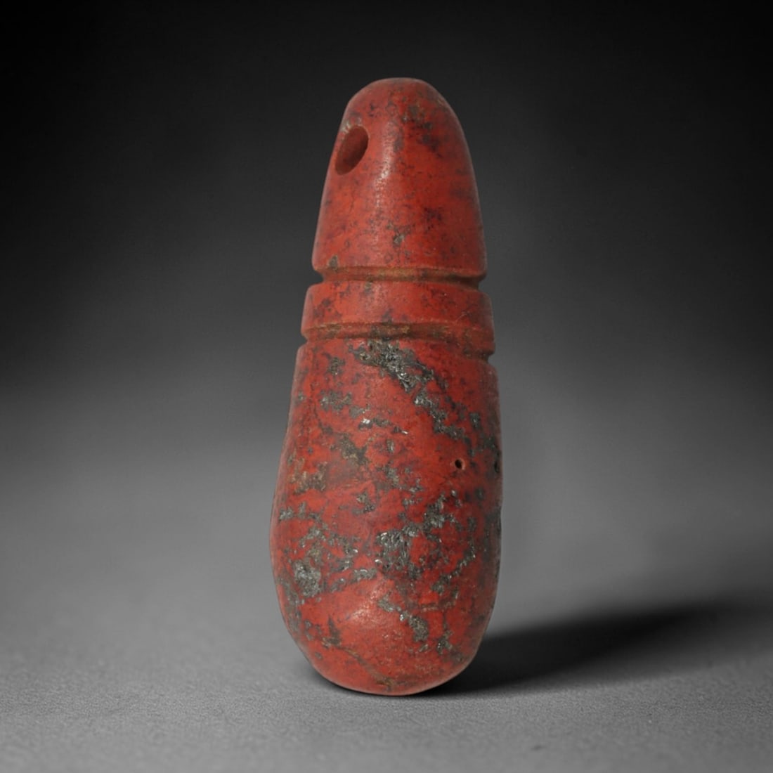 Ancient Red Stone Segmented Pendant - Near Eastern Ritual Amulet, 87XP: Carved from a rich, earthy red stone with natural dark mineral inclusions, this elongated pendant features a highly stylized segmented form, a distinctive geometric motif in ancient Near Eastern ritua