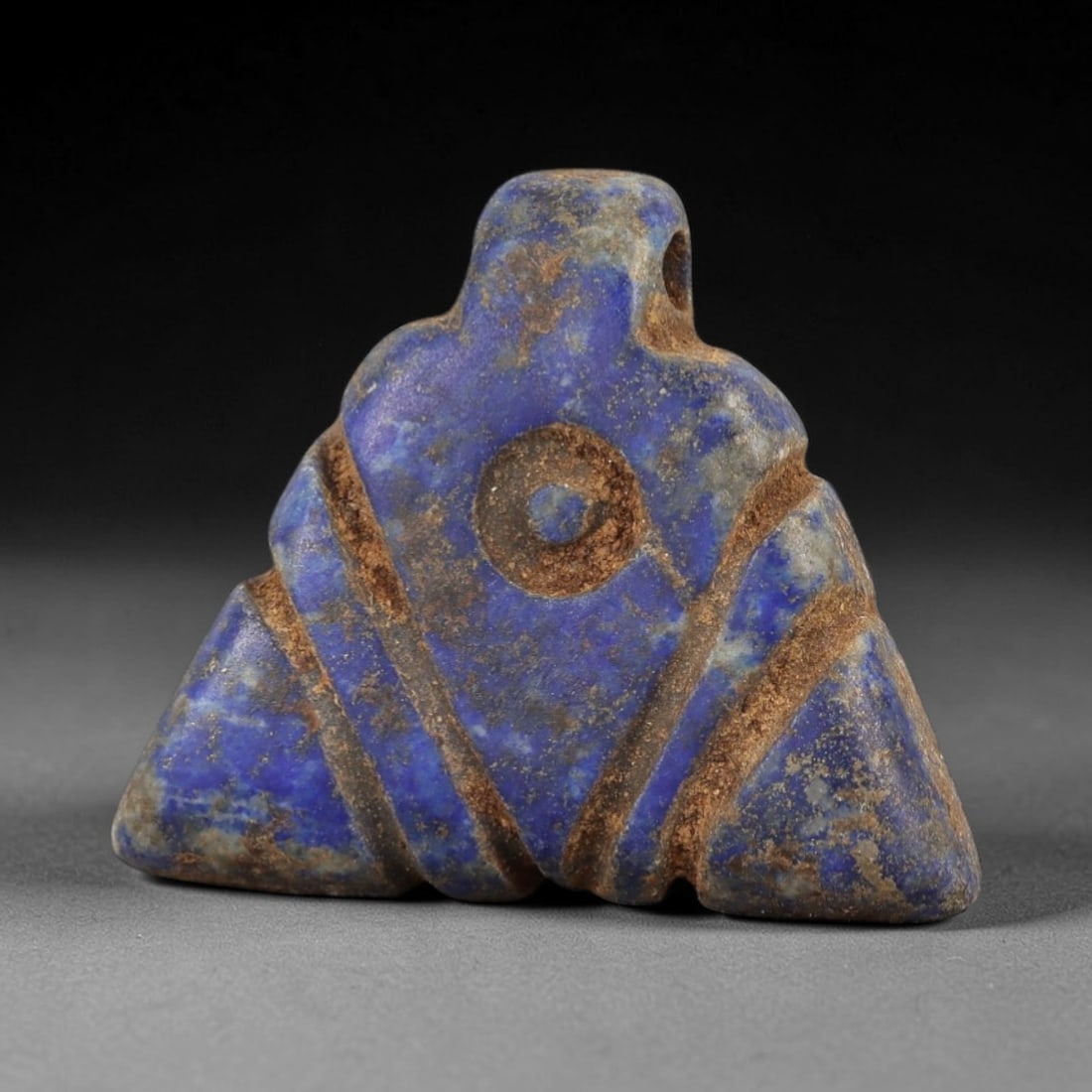 Ancient Lapis Lazuli Amulet - Egyptian Eye Symbol Pendant, 28AP: Carved from lapis lazuli, this amulet features a triangular form with a raised central circular eye motif, flanked by incised diagonal bands, with a drilled suspension hole piercing the upper lug. The