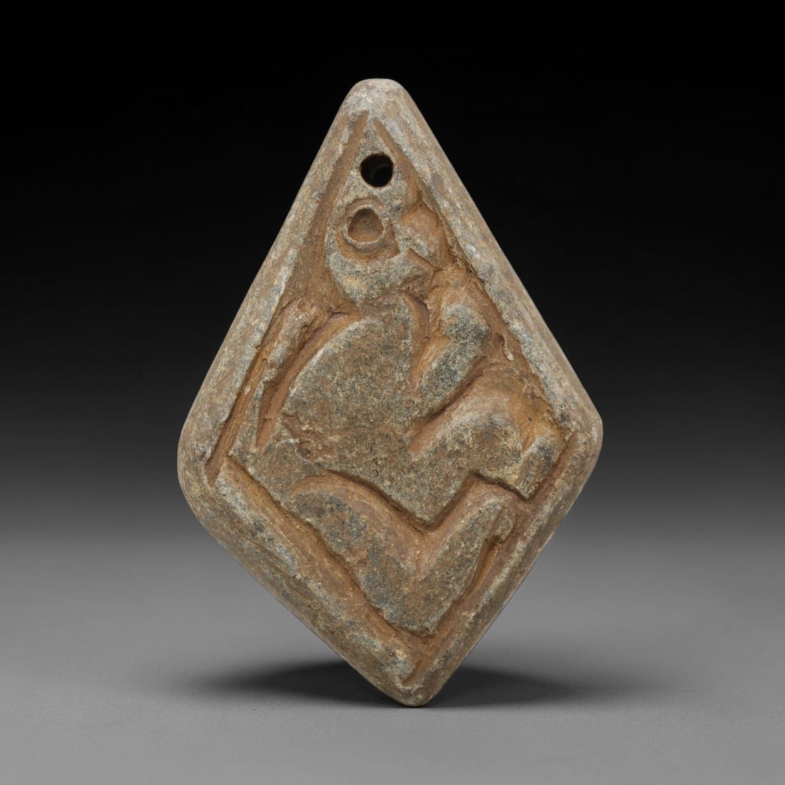 Ancient Schist Amulet - Near Eastern Carved Figurine, 57ZX: Carved from schist, the diamond-shaped amulet features a relief-carved stylized human figure in a crouched or seated pose, rendered with simplified anatomical details. A suspension hole is drilled thr