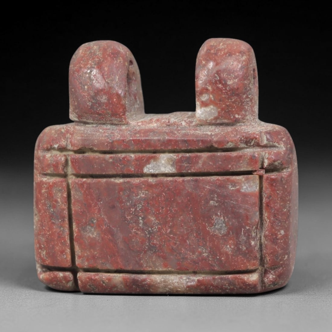Ancient Stone Double-Headed Amulet - Near Eastern Figurine, 69AP: Carved from a rich, reddish stone with natural mineral inclusions and weathered patina, this amulet features a rectangular, segmented body topped with two rounded, stylized heads. Each head bears subt