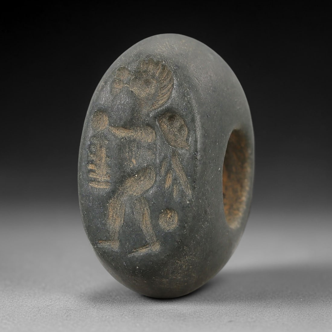 Ancient Steatite Stamp Seal - Near Eastern Figurine, 82AN: Carved from dark grey steatite, this hemispherical stamp seal features an incised anthropomorphic motif depicting a standing figure in profile, with distinct facial features, a stylized headdress, and