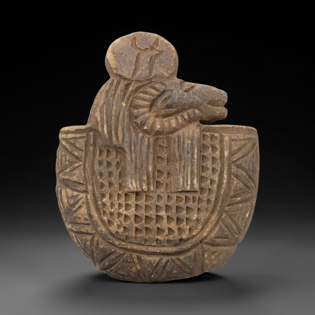 Ancient Stone Ram Amulet - Near Eastern Zoomorphic Figurine, 96WJ: Carved from a coarse, dark stone, this artifact takes the form of a stylized zoomorphic amulet featuring a ram's head surmounting a rounded, shield-shaped body. The ram is depicted with a prominent cu