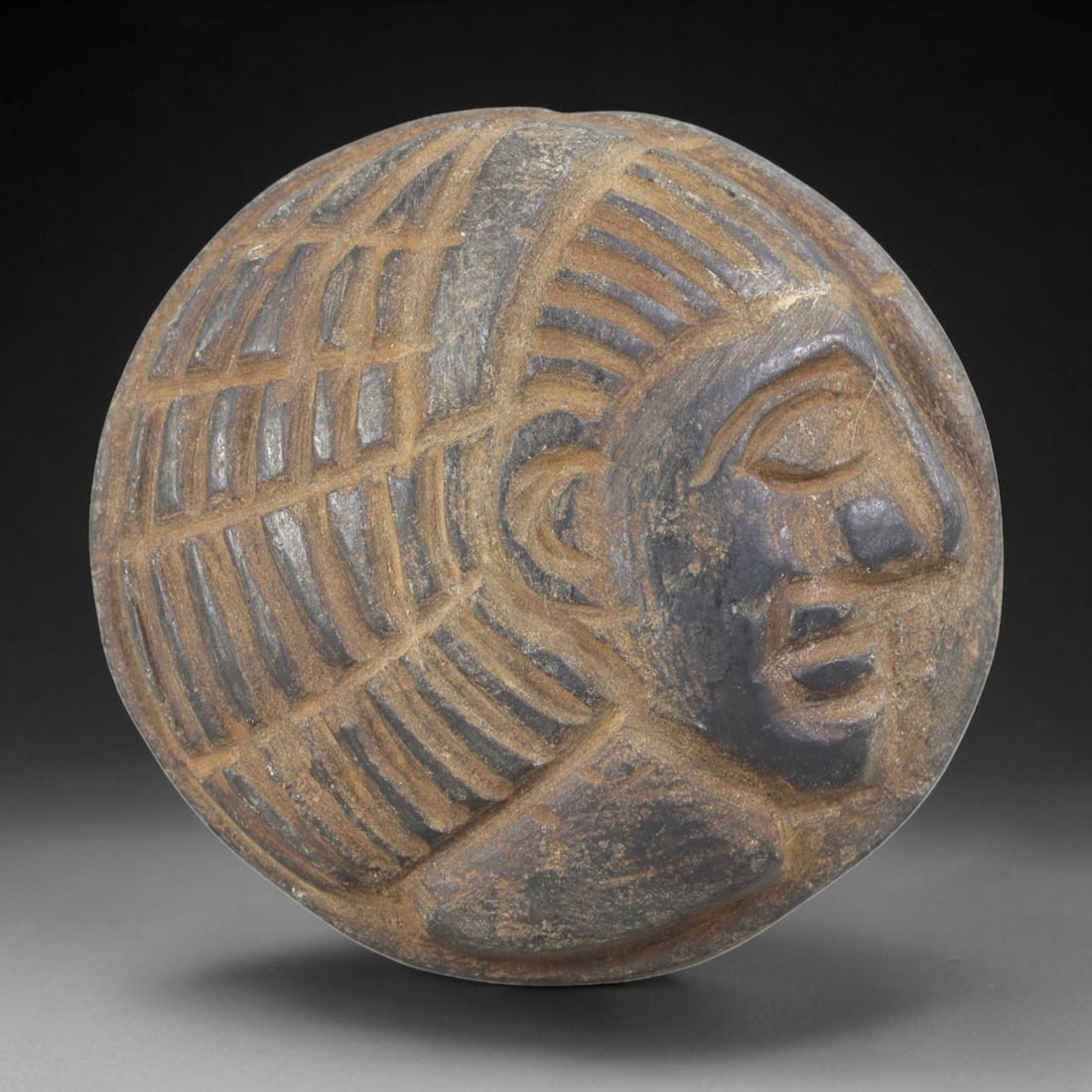 Ancient Stone Head Amulet - Near Eastern Bust Plaque, 44FY: Carved from a dark grey stone with warm brown mineral inclusions, this amulet takes the form of a human head bust in high relief, set within a circular plaque, reflecting the iconographic traditions o