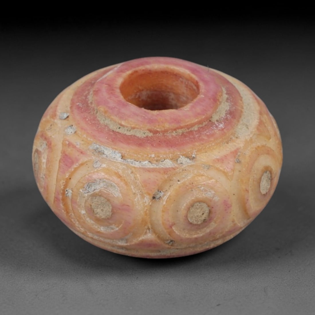 An Ancient Bone Spindle Whorl, 1st Millennium BCE, 72AC: Carved from pale, cream-colored bone with natural pinkish tonal variation, the spindle whorl is of a compressed, globular form, pierced with a central hole for attachment to a spindle shaft. The exter