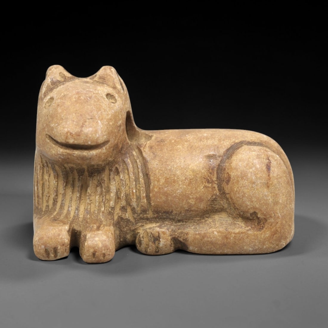 Ancient Stone Feline Figurine - Near Eastern Amulet, 4ZR: Carved from warm tan stone, this reclining feline figurine features a stylized head with incised eyes, pointed ears, and a gently curved mouth. The neck displays linear incised detailing suggesting a