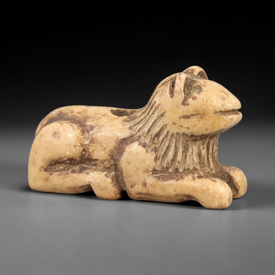 Ancient Stone Lion Figurine - Near Eastern Amulet, 85AL: Carved from light tan limestone, this amulet depicts a recumbent lion with a stylized mane rendered through incised vertical lines. The facial features are simplified with a single incised line for th