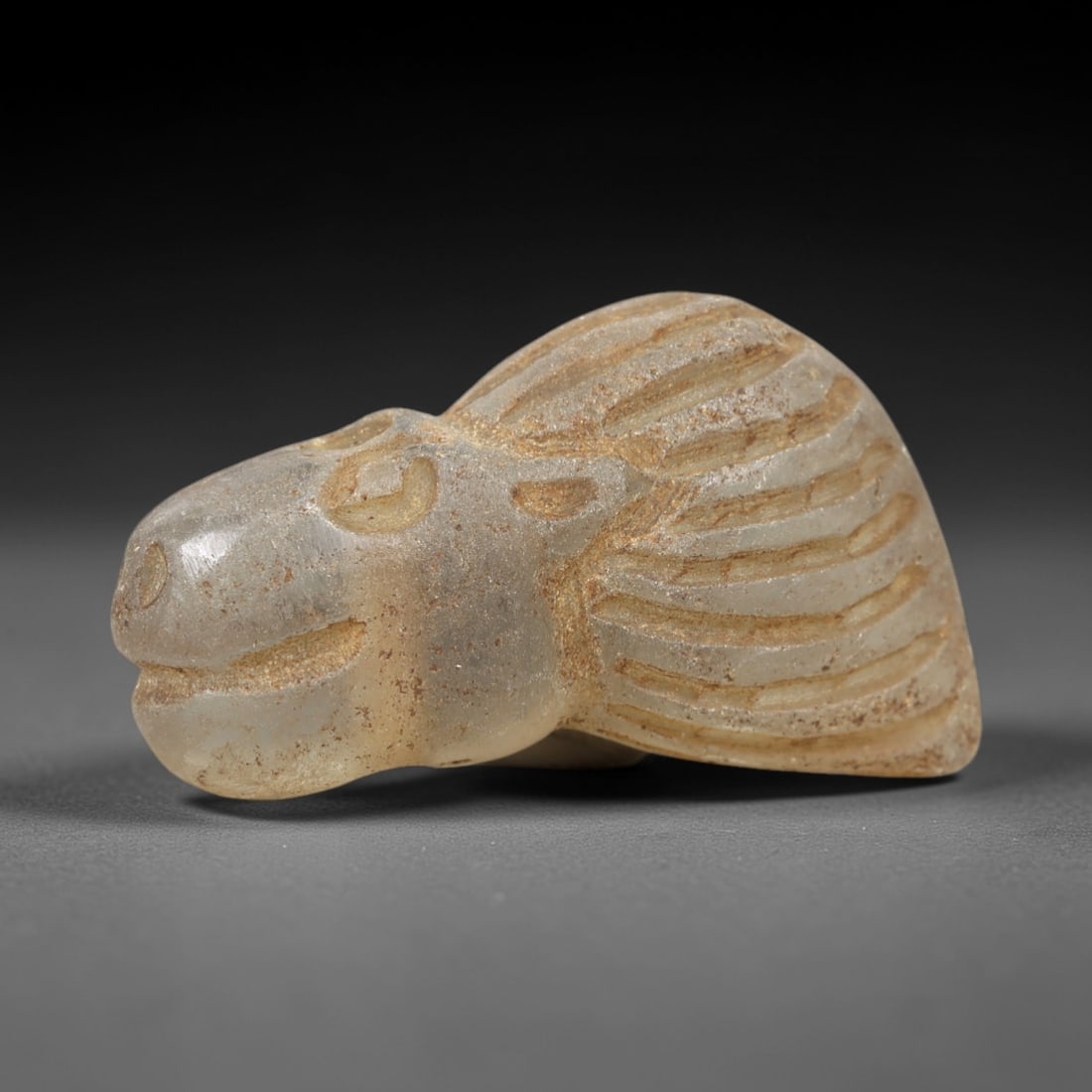 Ancient Glass Feline Head - Egyptian Zoomorphic Amulet, 23AL: This zoomorphic amulet is carved from translucent pale glass, shaped into the stylized head of a feline creature, likely a lioness or panther, with finely incised linear details defining the textured