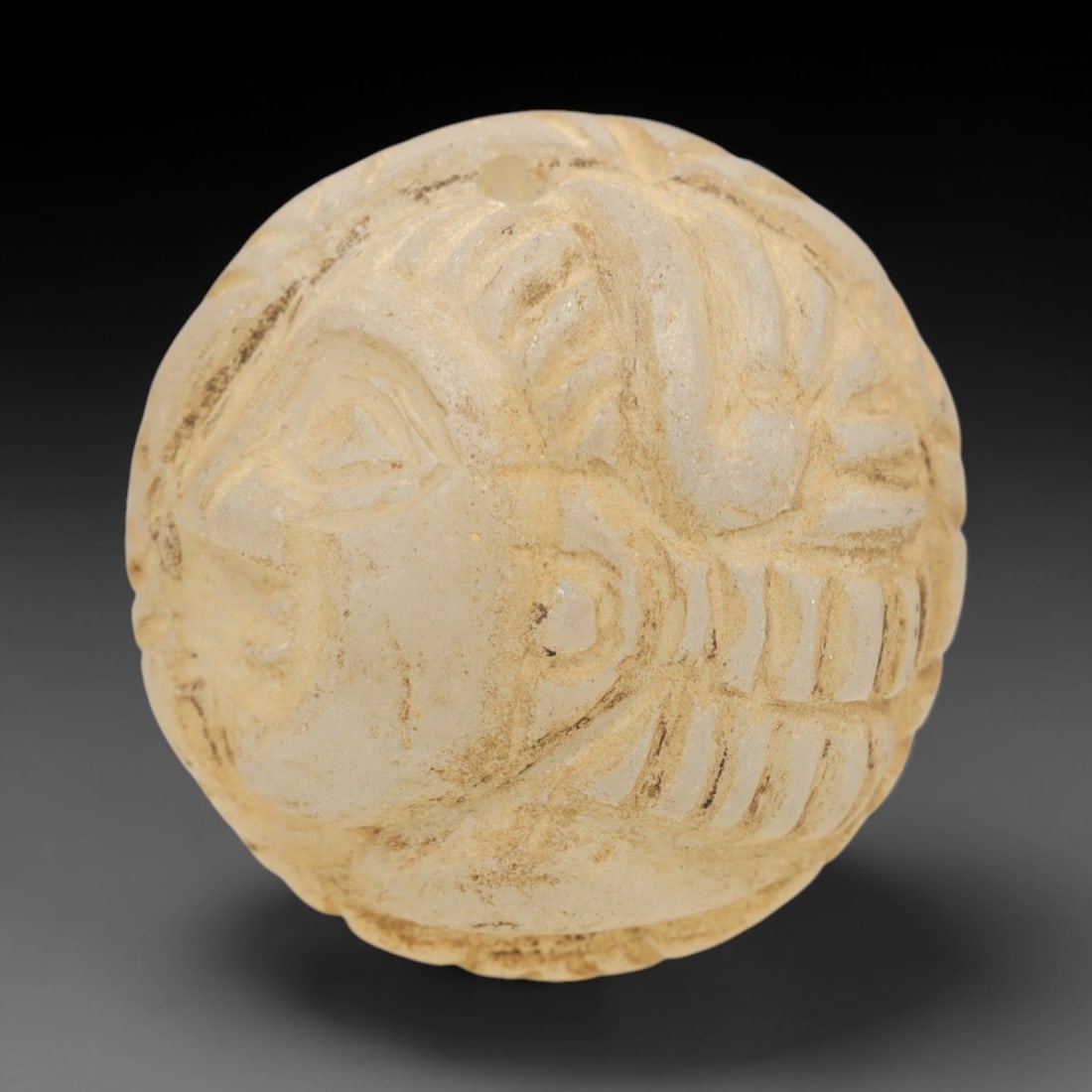 Ancient Glass Medieval Amulet - Near Eastern Bust Figure, 71ZR: Crafted from translucent pale glass with earthy encrustations, this spherical amulet features a raised relief profile bust of a human figure, with stylized linear details defining the hair, facial fea