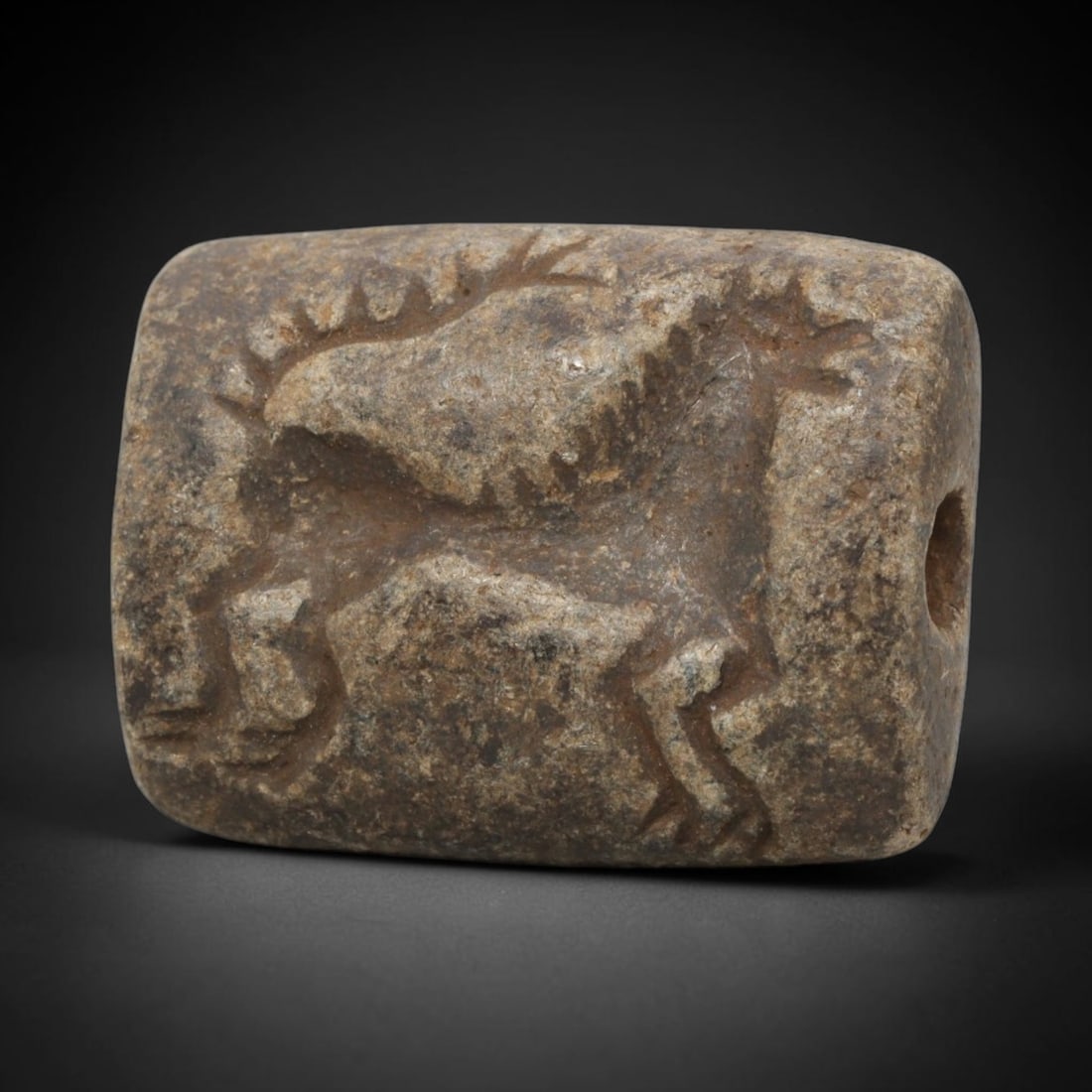 Ancient Stone Stamp Seal - Near Eastern Zoomorphic Relief, 28WJ: Carved from a dense, coarse stone, this artifact takes the form of a double convex stamp seal, adorned with a deeply incised zoomorphic relief motif. The central design depicts a stylized horned anima