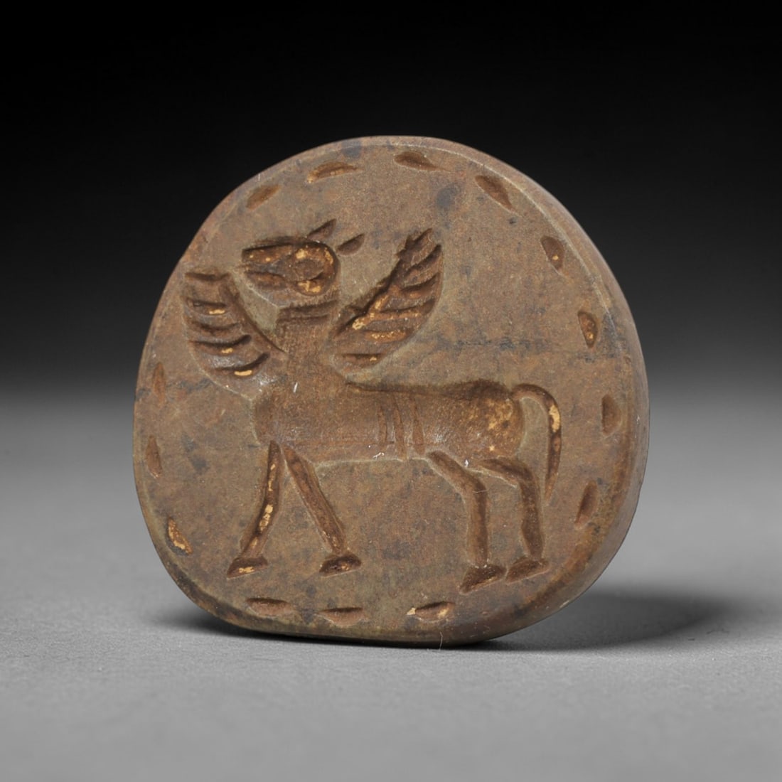 Ancient Steatite Stamp Seal - Near Eastern Winged Beast Figurine, 94ZV: This ancient Near Eastern stamp seal is carved from steatite, formed as a circular disc with incised decoration depicting a winged quadrupedal beast in profile, rendered with distinct linear detailing
