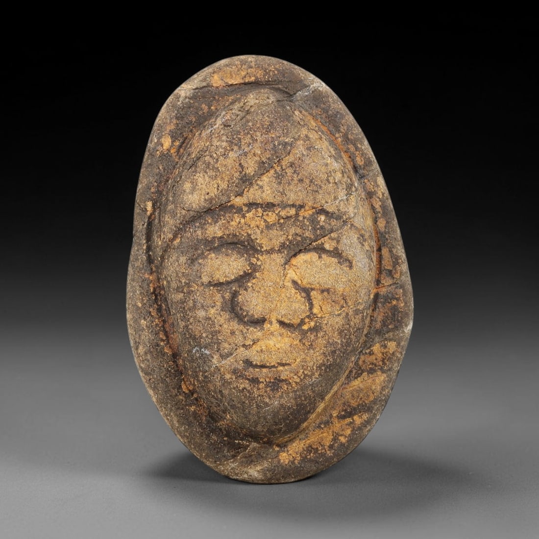 Ancient Stone Maskette - Near Eastern Amulet Figurine, 54WJ: Carved from stone, this ancient maskette features a stylized human face with incised carvings defining the closed eyes, nose, mouth, and a raised border framing the visage. The piece reflects the ritu