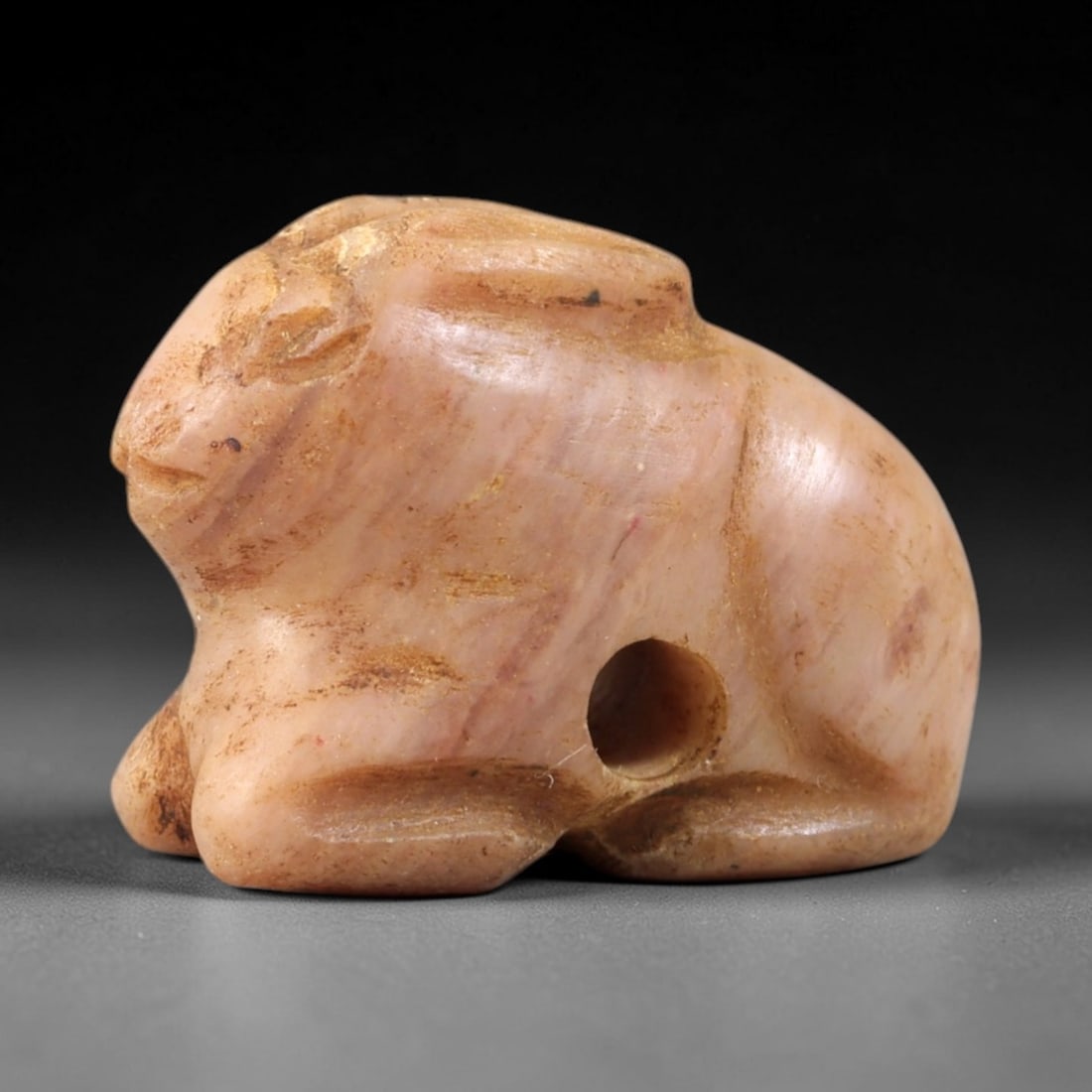 Ancient Pink Stone Hare - Near Eastern Amulet Figurine, 34AS: This amulet is carved from smooth pinkish stone, exhibiting a subtle, striated texture and warm earthy patina consistent with ancient Near Eastern burial contexts. The form depicts a recumbent hare, r