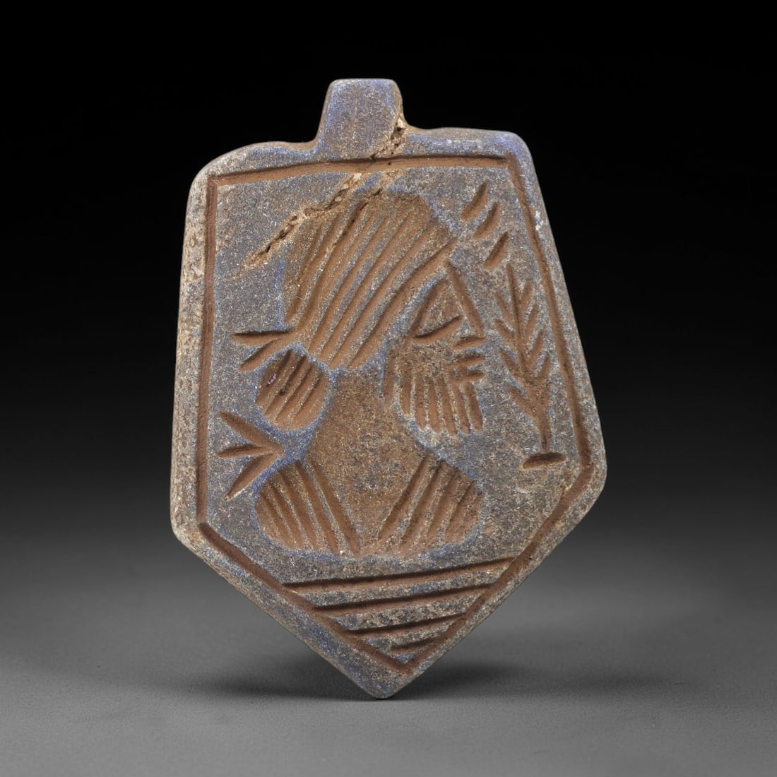 Ancient Lapis Lazuli Amulet - Near Eastern Profile Relief, 18FX: Carved from lapis lazuli, this shield-shaped amulet features a low-relief profile of a bearded male figure, with incised linear details defining the hairstyle, facial features, and upper body, alongsi