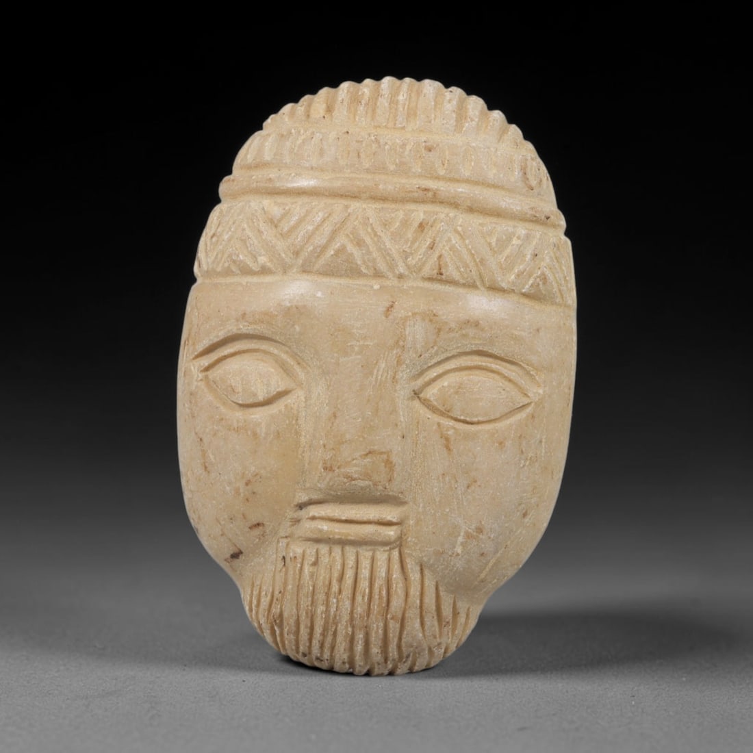 Ancient Stone Amulet - Near Eastern Bearded Male Mask Pendant, 77FY: Carved from light-colored stone, the amulet is an oval plaque crafted in the ancient Near Eastern style, featuring a raised relief portrait of a bearded male mask in frontal view. The surface bears in