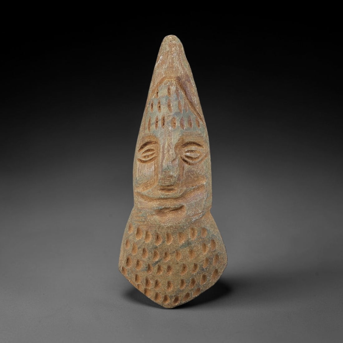 Ancient Stone Human Head Amulet - Near Eastern Carved Pendant, 50BA: Carved from hard stone (likely steatite or schist), this amulet takes the form of a stylized, three-dimensional human head in relief, rendered with detailed, stylized carving. The figure wears a tall,