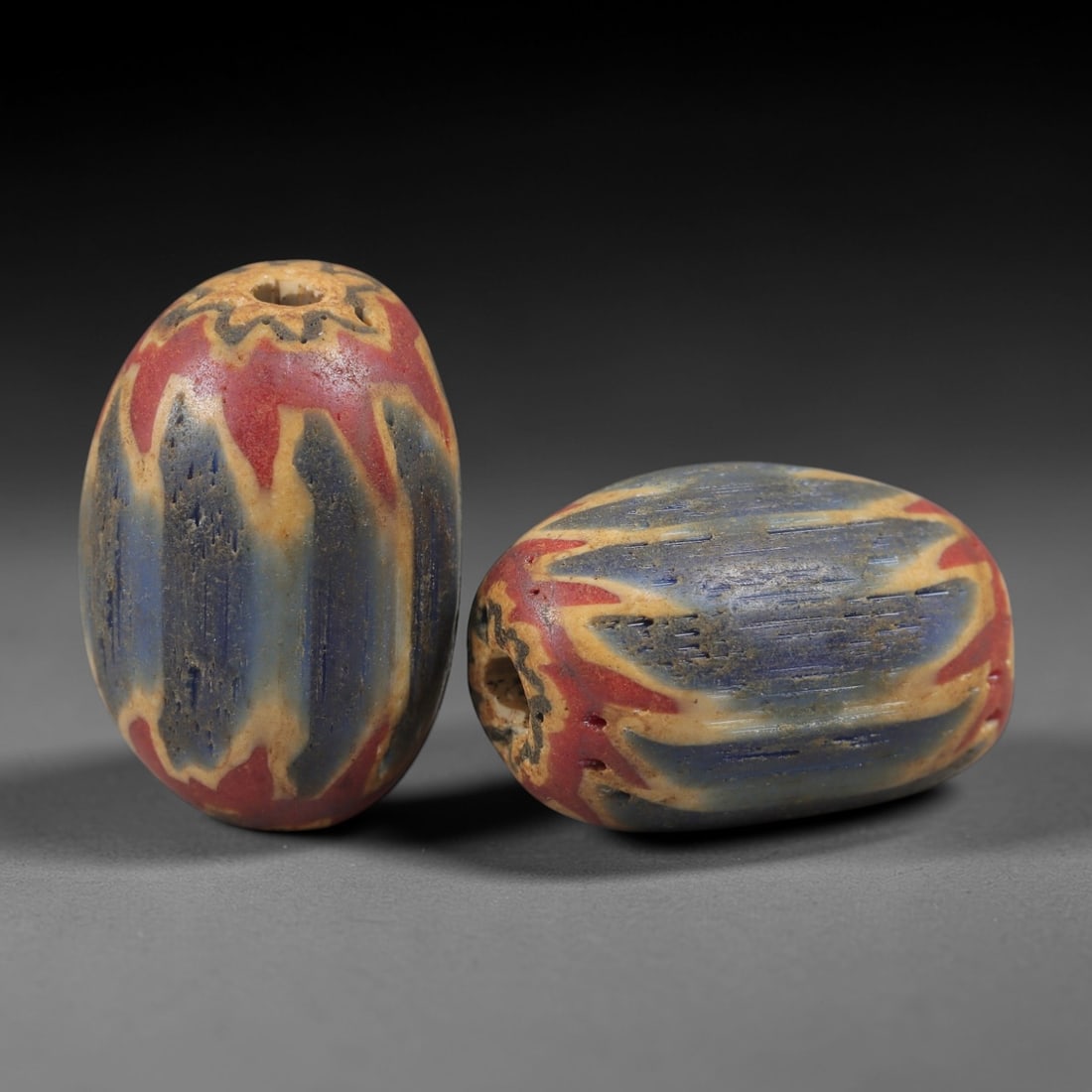 A Pair of Venetian Glass Chevron Beads, 18th-19th Century, 79AC: Crafted from translucent glass with a pale yellow core, each bead is decorated with a chevron pattern of alternating deep blue and vibrant red layers, radiating from the central perforation to the out