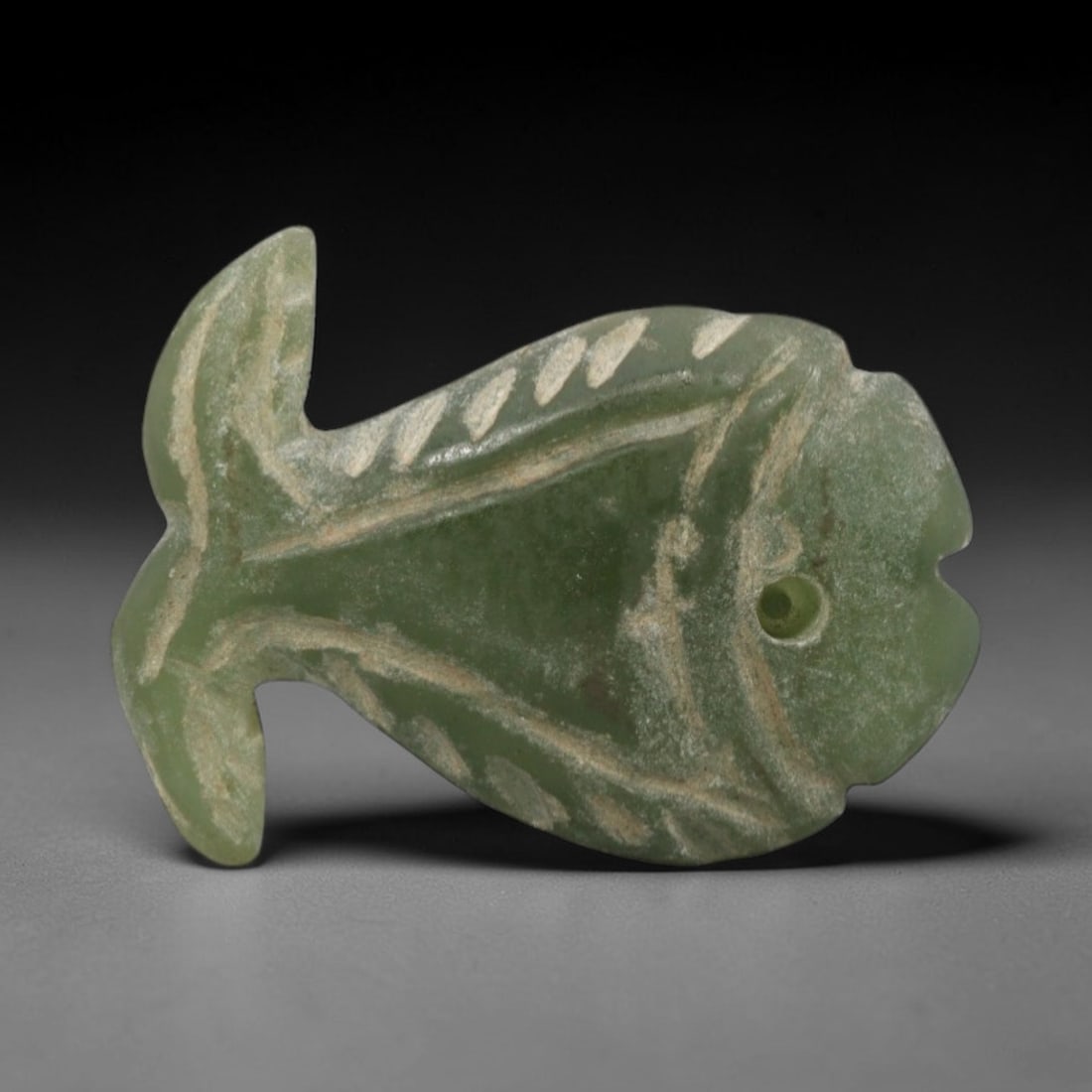 Ancient Egyptian Serpentine Fish Amulet - Carved Pendant Figure, 87ZW: This amulet is carved from a lustrous, pale green serpentine stone, with incised linear details defining its stylized fish form. The piece features a distinct head with an open mouth and a drilled sus