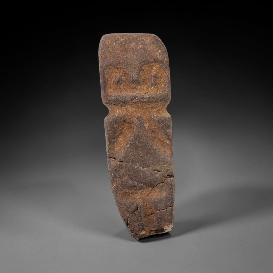 An Ancient Stone Idol, 20th Century, 64FW: Carved from a dark brown, porous stone with a naturally aged, patinated surface and subtle ochre inclusions, the idol is rectangular in form, with a stylized head featuring incised eyes and a nose, an