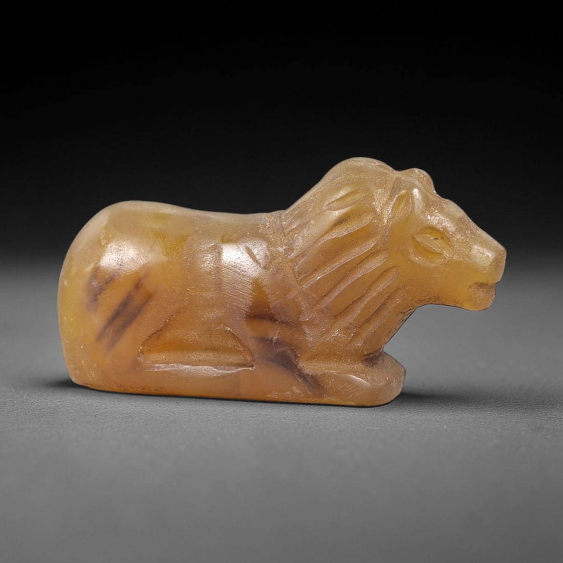 Ancient Agate Bovid Figurine - Near Eastern Amulet, 68AK: Carved from translucent golden-brown agate, this reclining bovid figurine features stylized curved horns, incised eyes, and linear detailing defining its mane. The compact, rounded body is polished sm