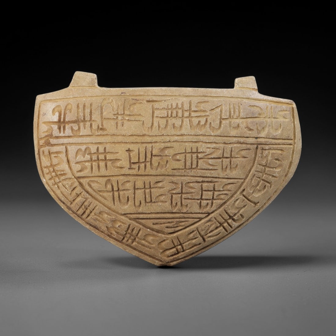 Ancient Stone Islamic Amulet - Carved Relic, 73BD: Carved from a dense, beige-hued stone, this artifact takes the form of a shield-shaped amulet, adorned with incised Islamic calligraphic inscriptions arranged in horizontal registers. The upper sectio