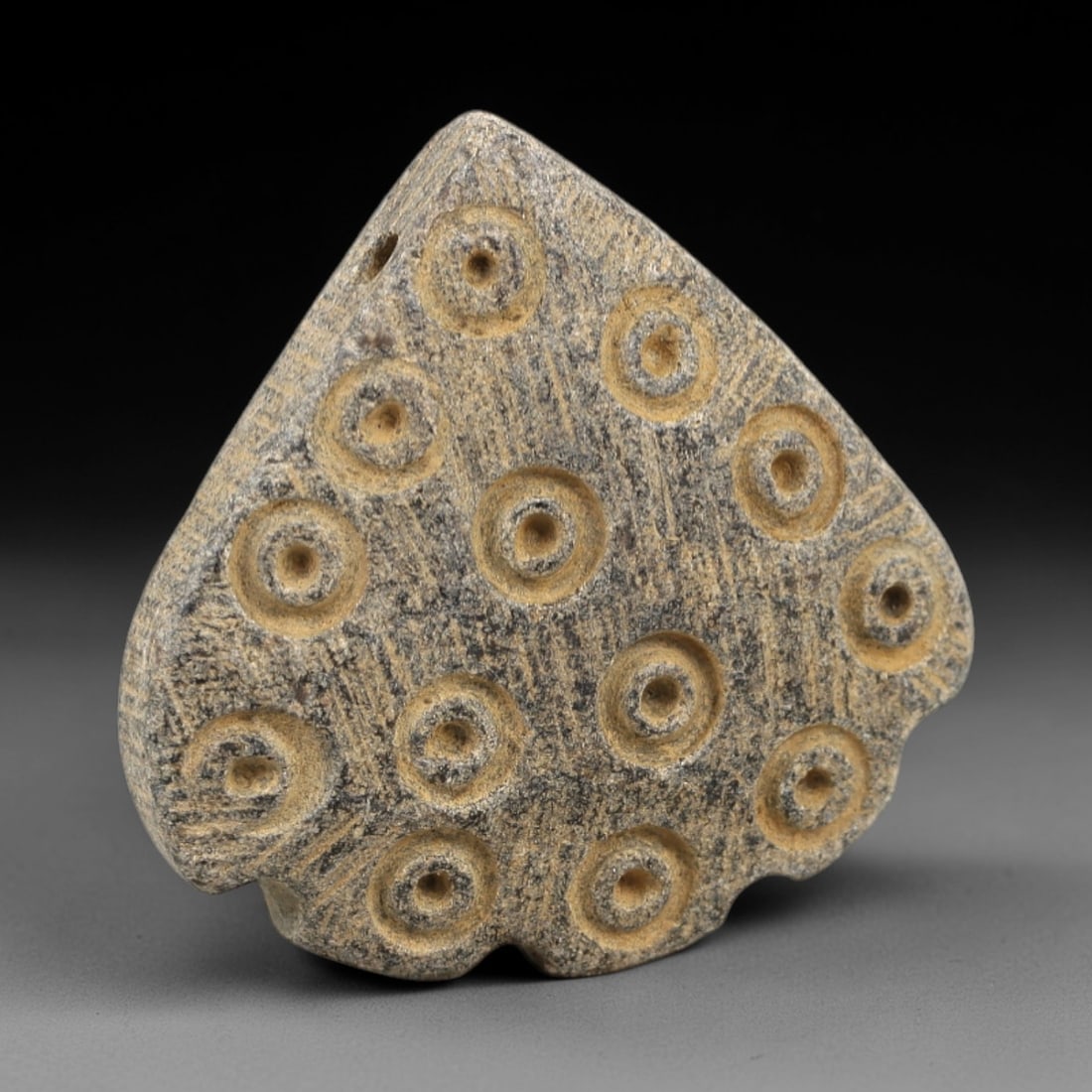 Ancient Stone Amulet - Near Eastern Concentric Circles, 7AL: This amulet is carved from granular stone, shaped into a heart-like form with a suspension hole at the upper edge, confirming its function as a wearable talisman. The surface is decorated with multipl
